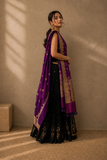 A woman models a traditional Indian outfit featuring a black fluted lehenga choli with gold embroidery, paired with a rich purple dupatta draped over her shoulder, set against a textured beige wall with warm sunlight shadows.