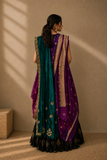 A woman stands with her back to the camera wearing a traditional Indian outfit consisting of a teal green and purple lehenga with gold zari embroidery and a matching dupatta, set against a warm beige wall with soft window shadows.