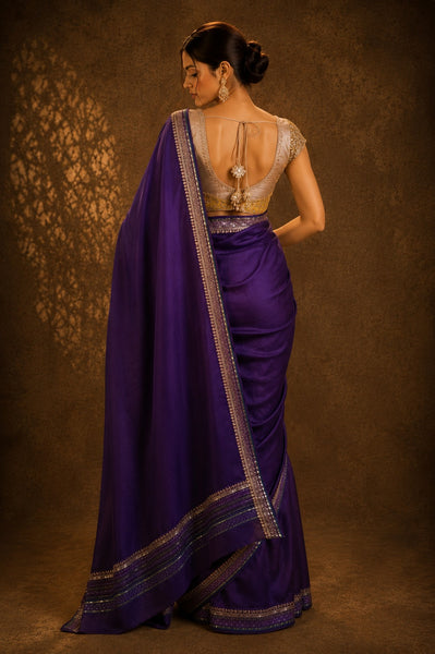 Woman wearing a purple saree with a gold blouse against a brown background