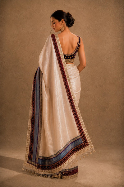 Woman wearing a traditional white saree with a brown and blue border against a beige background