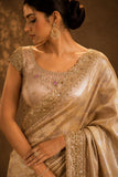 Woman wearing a beige saree with intricate embroidery against a brown background