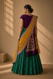 A woman wearing a traditional Indian outfit with a deep purple short-sleeve blouse, a green pleated skirt, and a mustard yellow dupatta with intricate gold and silver embroidery, standing in a warm-lit room.
