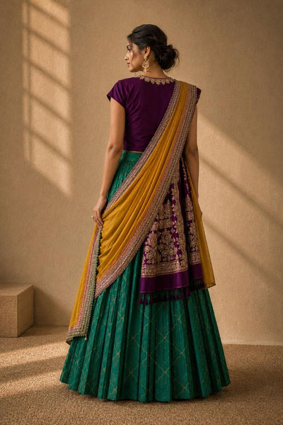 A woman wearing a traditional Indian outfit with a deep purple short-sleeve blouse, a green pleated skirt, and a mustard yellow dupatta with intricate gold and silver embroidery, standing in a warm-lit room.