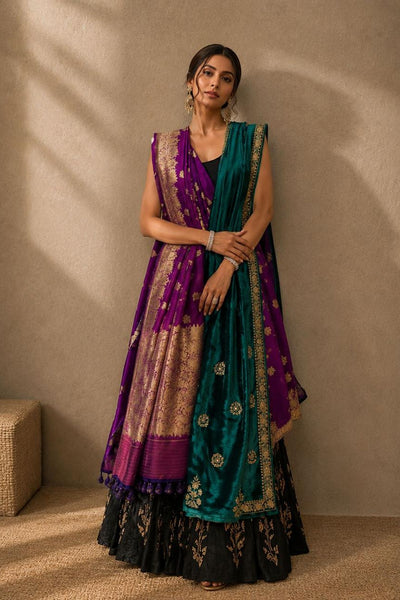 A woman models a traditional Indian outfit consisting of a black flared skirt with gold embroidery, a purple sleeveless vest, and a teal green dupatta with gold motifs, set against a textured beige wall with soft shadows.