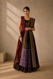 A woman models a traditional Indian outfit consisting of a sleeveless maroon and black embroidered vest, a matching flared skirt with purple and gold patterns, and a draped dupatta, set against a textured beige wall with warm sunlight shadows.