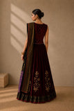 A woman stands with her back to the camera wearing a deep maroon velvet traditional outfit with gold embroidery and a patterned dupatta, set against a warm beige textured wall with soft window shadows.