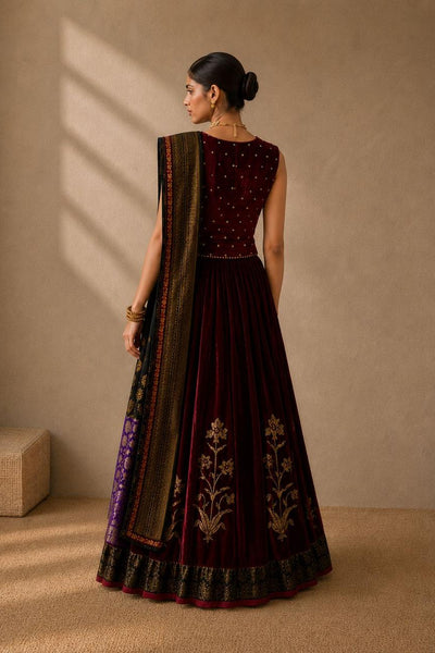 A woman stands with her back to the camera wearing a deep maroon velvet traditional outfit with gold embroidery and a patterned dupatta, set against a warm beige textured wall with soft window shadows.