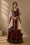 A woman models a traditional Indian outfit consisting of a deep maroon velvet skirt with gold embroidery, a matching embroidered sleeveless top, and a cream-colored dupatta draped over her shoulders, set against a textured beige wall with soft window shadows.