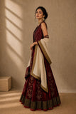 A woman models a traditional Indian outfit consisting of a deep maroon lehenga choli with gold embroidery, paired with a cream dupatta featuring gold borders, set against a warm beige textured wall with soft window shadows.