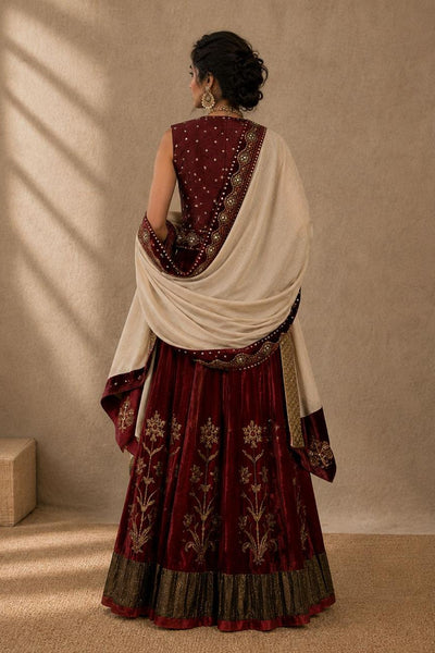 A woman shown from the back wearing a traditional Indian outfit with a deep maroon skirt decorated with gold embroidery, a matching embroidered sleeveless top, and a draped off-white dupatta, set against a textured beige wall with soft window shadows.