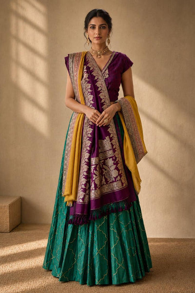 A woman models a traditional Indian ethnic outfit consisting of a deep purple embroidered kurta, a green pleated skirt, and a yellow and purple patterned dupatta, set against a warm beige wall with soft window shadows.