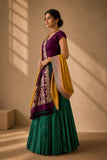 A woman models a traditional Indian outfit consisting of a deep green pleated skirt, a purple sleeveless blouse, and a yellow dupatta with intricate purple and gold patterns, set against a warm beige wall with window shadow details.