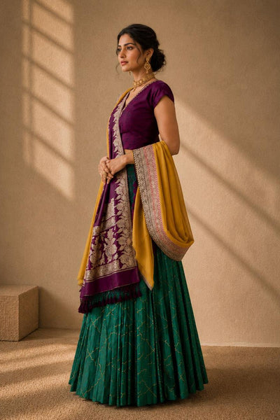 A woman models a traditional Indian outfit consisting of a deep green pleated skirt, a purple sleeveless blouse, and a yellow dupatta with intricate purple and gold patterns, set against a warm beige wall with window shadow details.