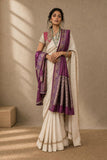 A woman models a traditional Indian saree with a white body, purple border, and a matching purple dupatta, standing in a warm-toned studio setting.