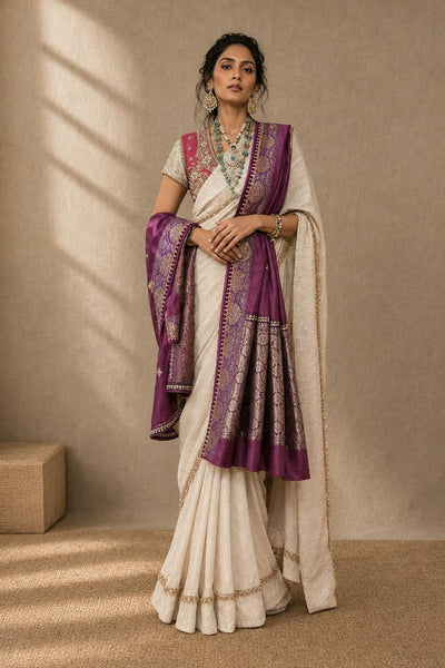 A woman models a traditional Indian saree with a white body, purple border, and a matching purple dupatta, standing in a warm-toned studio setting.