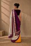 A woman stands with her back to the camera wearing a traditional Indian saree. The saree features a deep purple border and pallu, a white body with gold zari embroidery, and a contrasting yellow lower skirt.