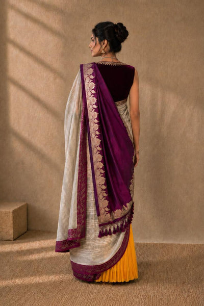 A woman stands with her back to the camera wearing a traditional Indian saree. The saree features a deep purple border and pallu, a white body with gold zari embroidery, and a contrasting yellow lower skirt.