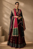 A woman models a traditional Indian outfit consisting of a deep maroon velvet skirt with gold embroidery, a black embroidered kurta, and a pink dupatta with green and gold borders, set against a warm beige background with soft shadows.
