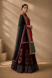 A woman models a traditional Indian outfit consisting of a deep maroon velvet skirt with gold floral embroidery, a black embroidered blouse, and a black dupatta draped over her shoulders, set against a warm beige textured wall with soft window shadows.