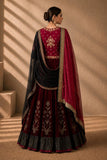 A woman wearing a traditional Indian outfit with a deep maroon flared skirt, a matching embroidered blouse, and a sheer black dupatta draped over her shoulders, photographed in a warm indoor setting with sunlight and shadows.