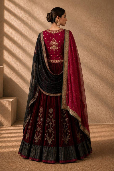 A woman wearing a traditional Indian outfit with a deep maroon flared skirt, a matching embroidered blouse, and a sheer black dupatta draped over her shoulders, photographed in a warm indoor setting with sunlight and shadows.