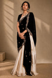 A woman models a traditional black and ivory saree with intricate silver embroidery, set against a warm beige textured wall with soft shadow patterns.