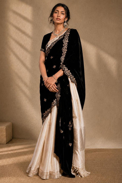 A woman models a traditional black and ivory saree with intricate silver embroidery, set against a warm beige textured wall with soft shadow patterns.