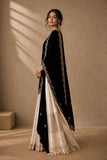 A woman models a traditional black velvet anarkali-style dress with gold embroidery and a cream-colored skirt, set against a textured beige wall with soft window shadow light.