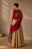 A woman wearing a traditional Indian outfit with a deep red embroidered sleeveless top, a beige flared skirt, and a red dupatta with a purple patterned border, shown from the back against a textured beige wall.