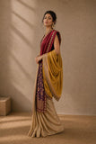 A woman models a traditional Indian outfit featuring a beige kurta, a deep red sleeveless overlay, and a mustard yellow dupatta with intricate gold embroidery, set against a textured beige wall with soft window shadows.