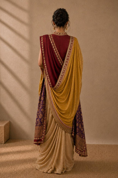 A woman stands with her back to the camera wearing a draped saree in mustard yellow, deep maroon, and beige with ornate gold and purple borders, set against a warm beige wall with soft window shadow patterns.