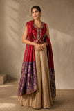 A woman models a traditional Indian outfit consisting of a sleeveless red embroidered vest, a beige pleated skirt, and a red dupatta with purple and gold border patterns, set against a textured beige studio backdrop.