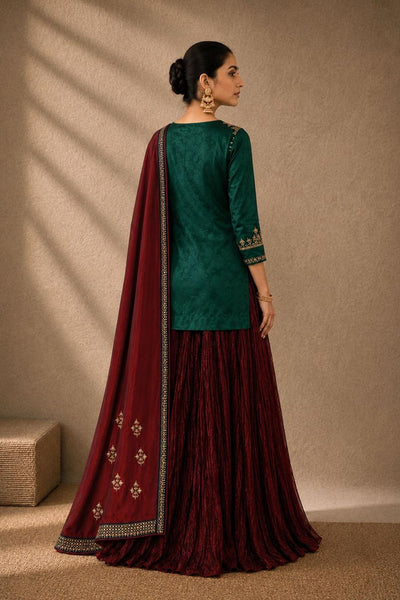 A woman wearing a traditional Indian outfit with a dark green velvet kurta, a pleated dark red skirt, and a matching dupatta with gold embroidery, standing in profile against a textured beige wall.