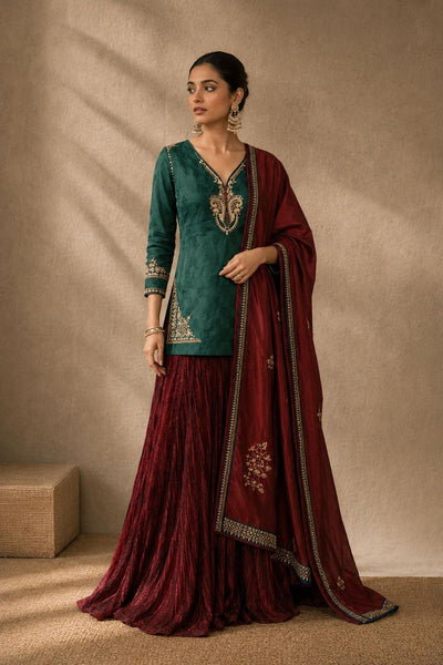 A woman models a traditional Indian outfit consisting of a teal green embroidered kurta, a deep maroon flared skirt, and a matching maroon dupatta with gold borders, set against a textured beige studio backdrop.