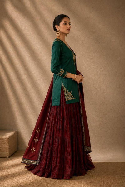 A woman models a traditional Indian outfit consisting of a dark green kurta with gold embroidery, paired with a deep maroon pleated skirt and a matching maroon dupatta draped over her shoulder, set against a warm beige background with soft window shadow patterns.