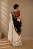 A woman stands with her back to the camera wearing a traditional Indian saree. The saree has a deep red embroidered blouse, a black drape, and a white body with gold border detailing.