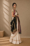 A woman models a traditional Indian outfit consisting of a cream-colored skirt, a pink and gold embroidered blouse, and a black dupatta with gold borders, set against a textured beige wall with soft window shadows.