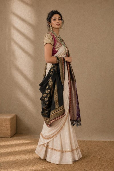 A woman models a traditional Indian outfit consisting of a cream-colored skirt, a pink and gold embroidered blouse, and a black dupatta with gold borders, set against a textured beige wall with soft window shadows.