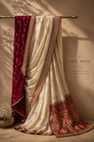 A draped cream-colored saree with a deep red border and a matching red embroidered pallu, hanging from a brass rod against a textured beige wall with decorative shadows. The brand name 'ANJU MODI' is printed on the wall.